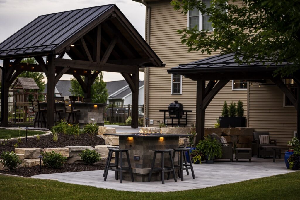Two timber pavilions with black metal roofs over a paver patio and stone fire bar in Neenah, Wisconsin
